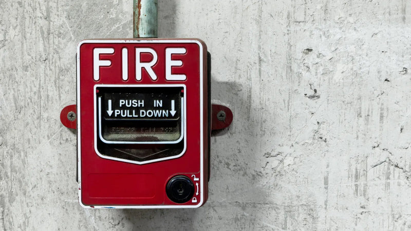 A vintage-style red fire alarm pull station mounted on a textured light gray concrete wall. The red box features white 'FIRE' lettering at the top, a black 'PUSH IN PULL DOWN' label in the center, and a small black switch at the bottom.
